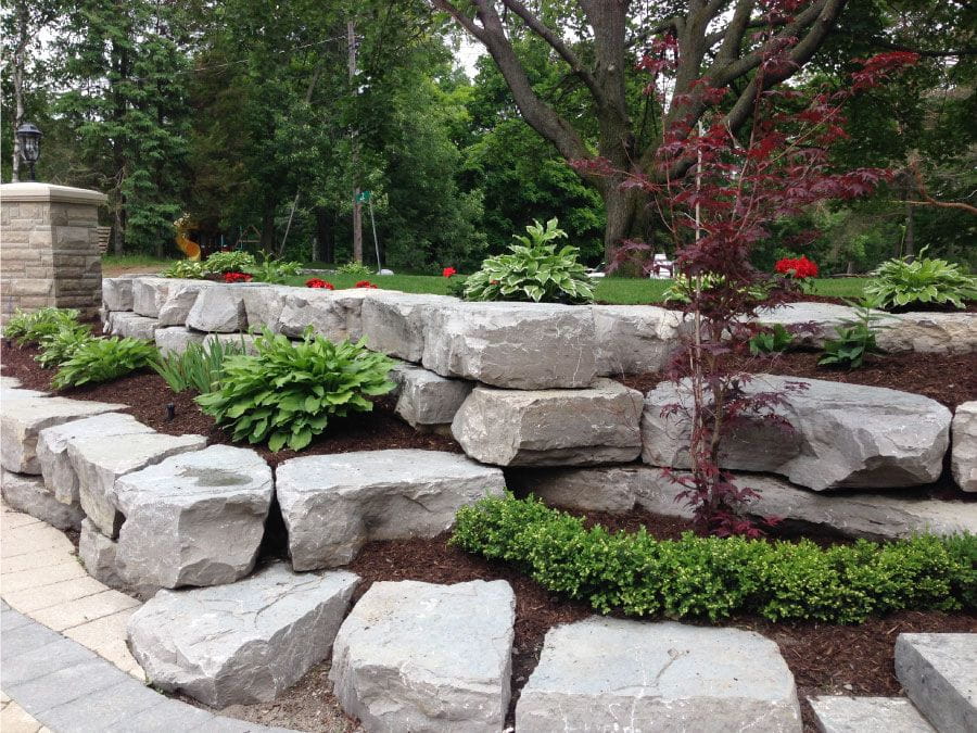 Stone retaining wall with planted beds, including green bushes and red flowers, with trees in the background.