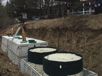 Man inspecting a septic system installation. Tanks sit in a trench, with two circular filter units in foreground.