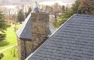 Slate roof and stone chimney on a building with a turret, set in a grassy area with a view of water and trees.