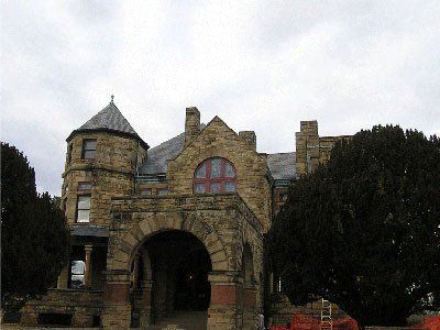 Stone building with arched entrance, tower, and dark trees against a cloudy sky.