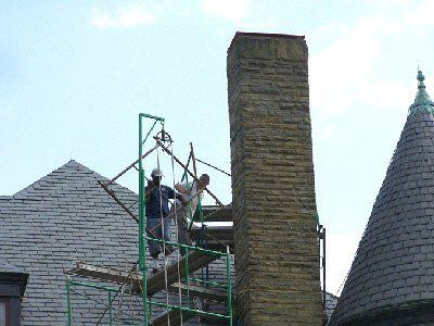 Two workers on a scaffold repair a stone chimney on a slate roof under a partly cloudy sky.