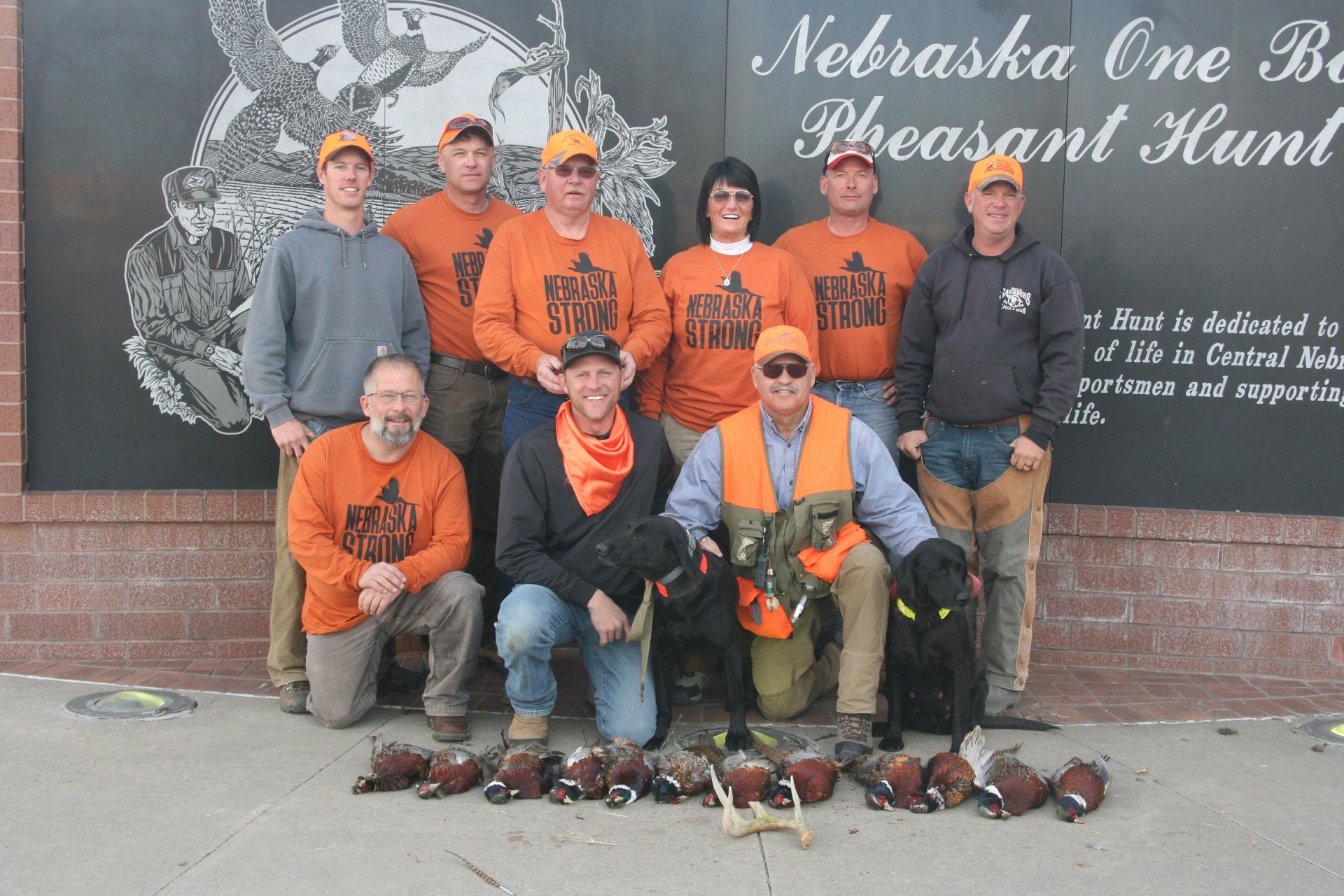 Group of hunters with orange shirts and hats pose with a dog and pheasants. 