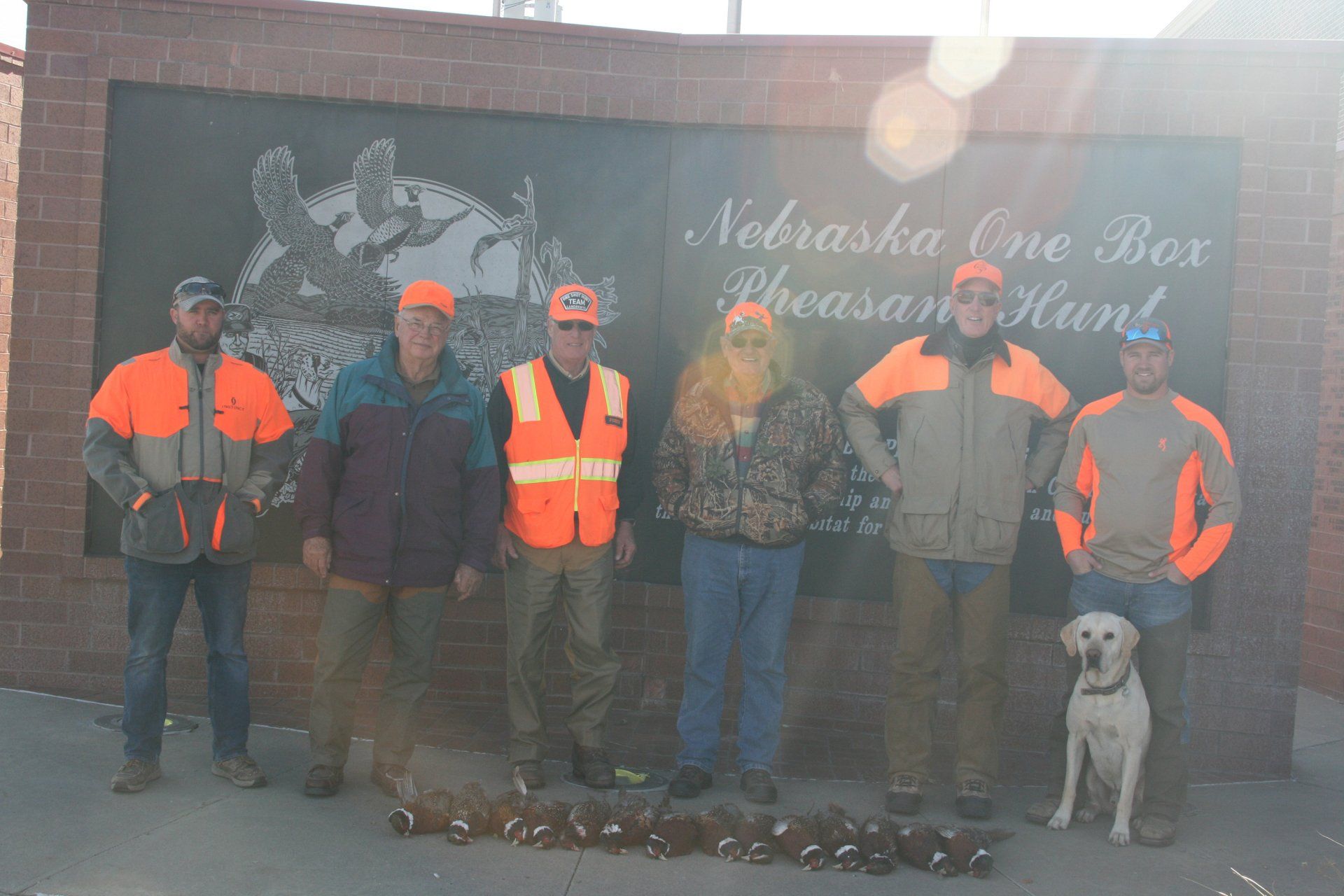 Group of six hunters with a dog posing by a sign after a pheasant hunt, sunny day.