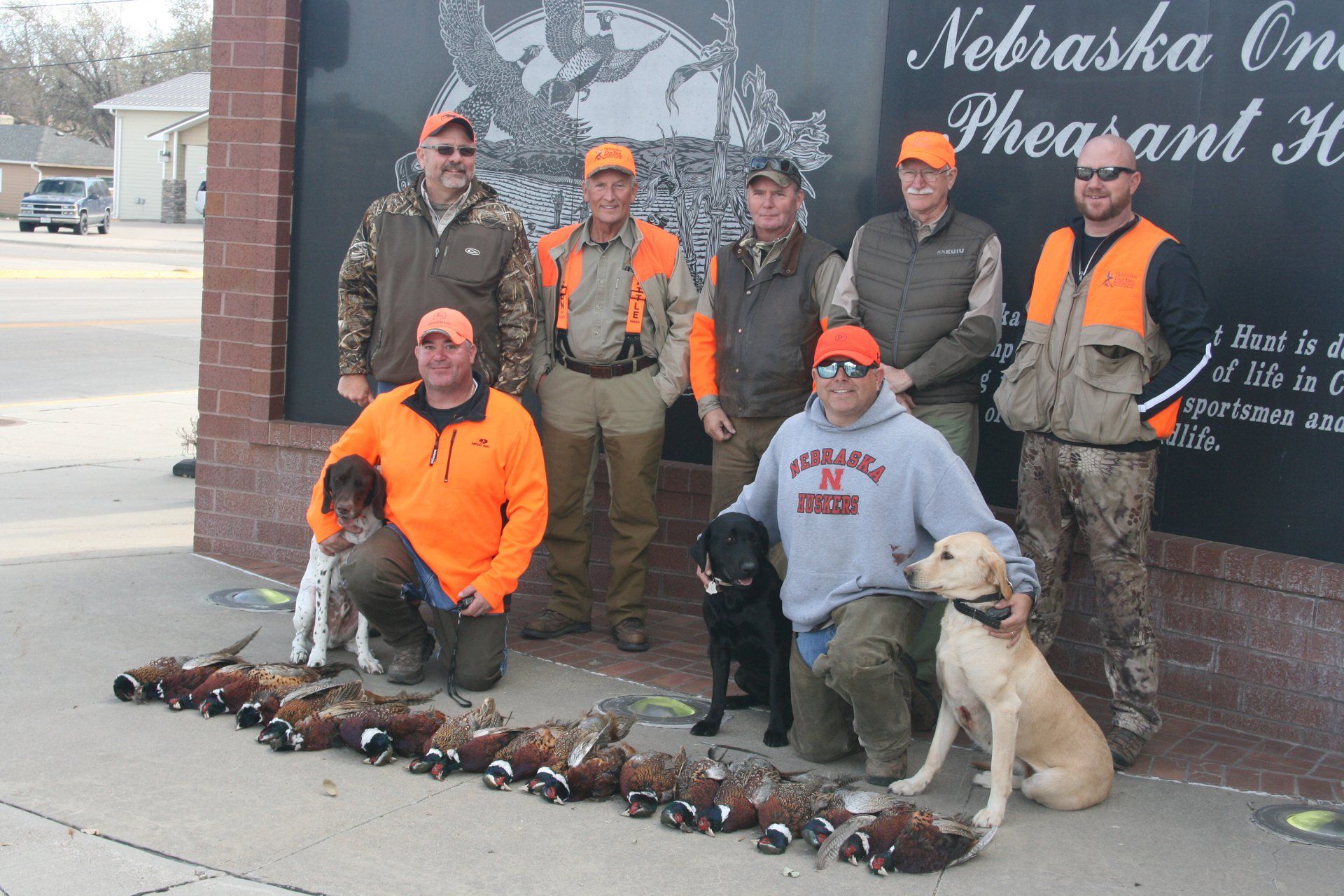 Group of hunters with dogs and harvested pheasants posing outside a building in Nebraska.