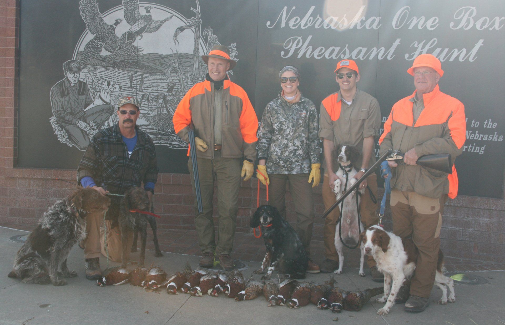 Five hunters with dogs and pheasants in front of a building with a sign.