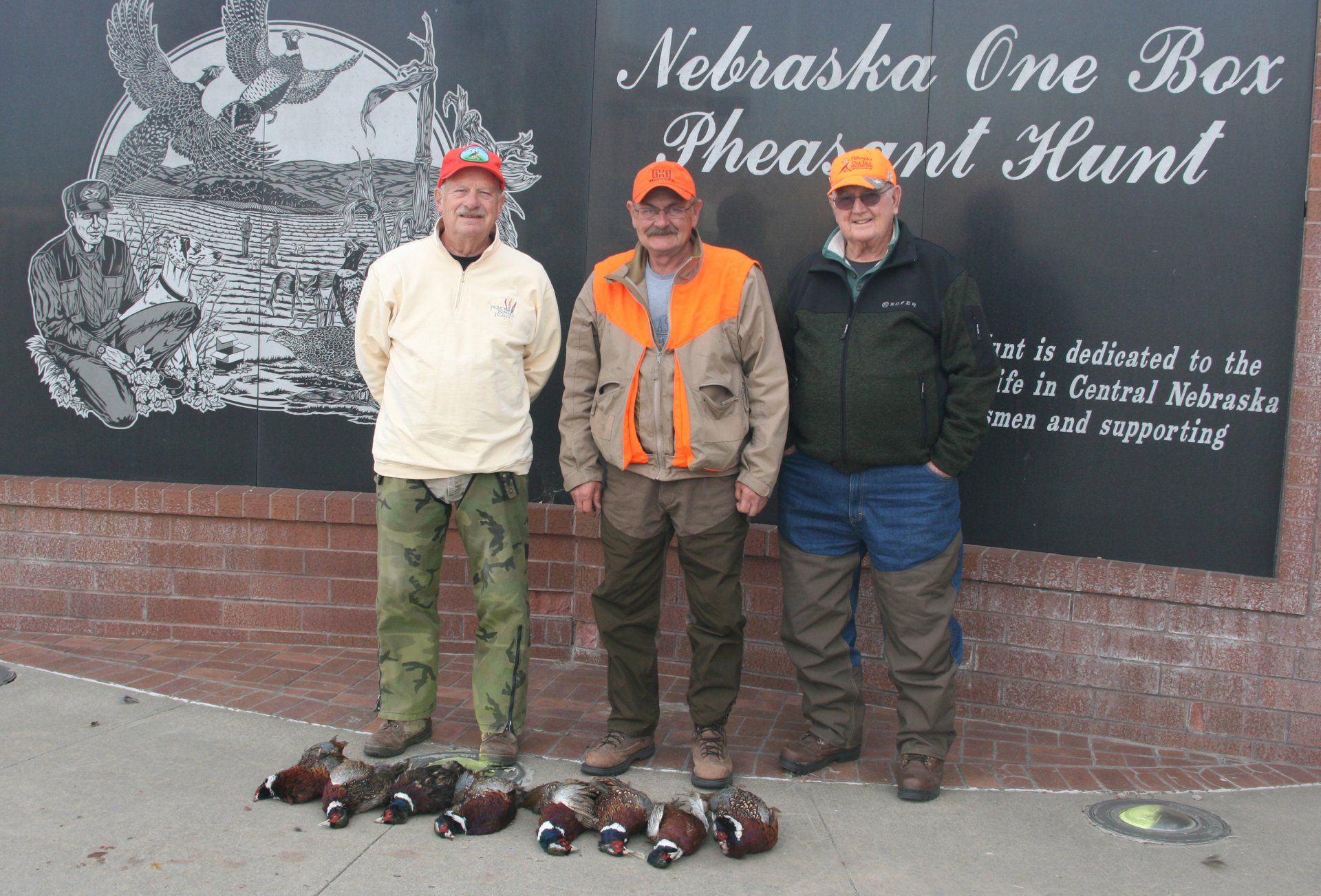 Three hunters stand with their pheasant haul in front of a sign for the Nebraska One Box Pheasant Hunt.