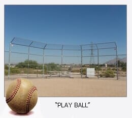 Baseball on the ground with a chain-link fence in background under a blue sky.