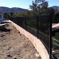 Black metal fence atop a concrete wall, with a dirt foreground and distant mountains.
