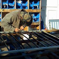 Welder wearing a helmet working on a metal gate outdoors, using a welding torch.