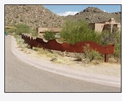 A rusty metal fence curves along a road in a desert landscape.