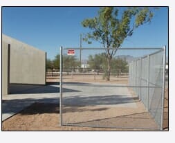 Chain-link fence in front of a building. Sandy ground, blue sky, and a tree in the background.