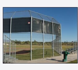 Baseball batting cage with metal frame, netting, and dark screen panels on a field under a clear blue sky.