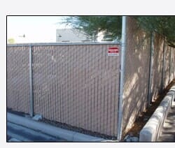 Chain-link fence with brown privacy slats; sign, sidewalk, and partial tree visible.