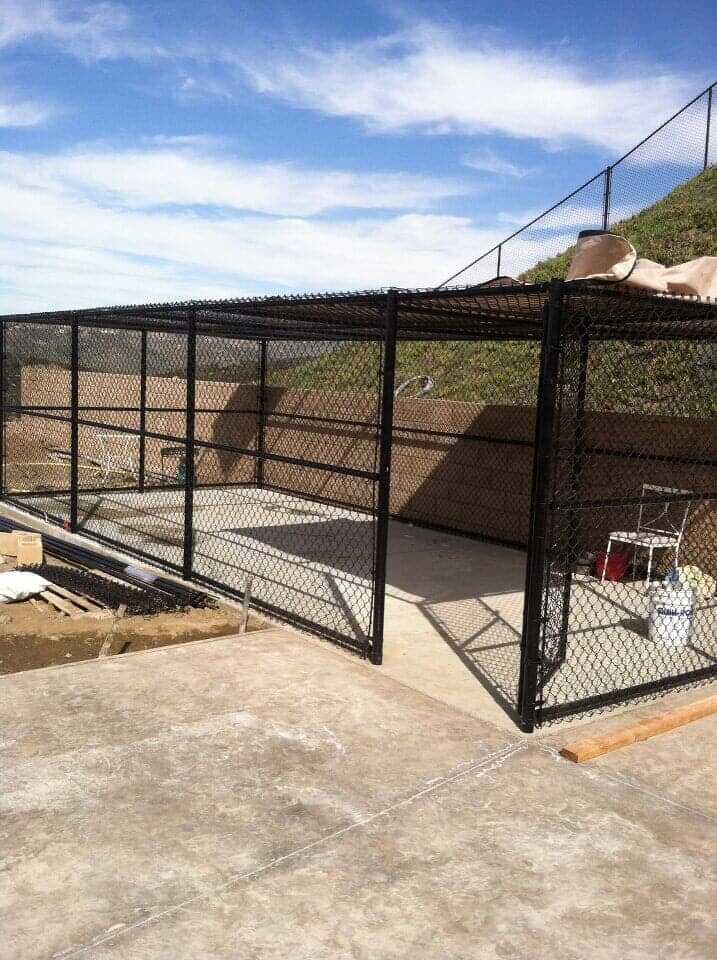 Black metal enclosure with chain link fencing on a concrete patio, against a grassy hill and blue sky.