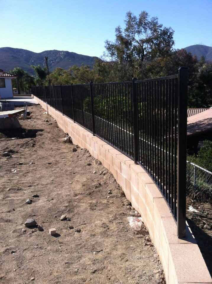 Black metal fence atop a curved, light-brown brick wall, in a dirt yard with a view of hills and trees.