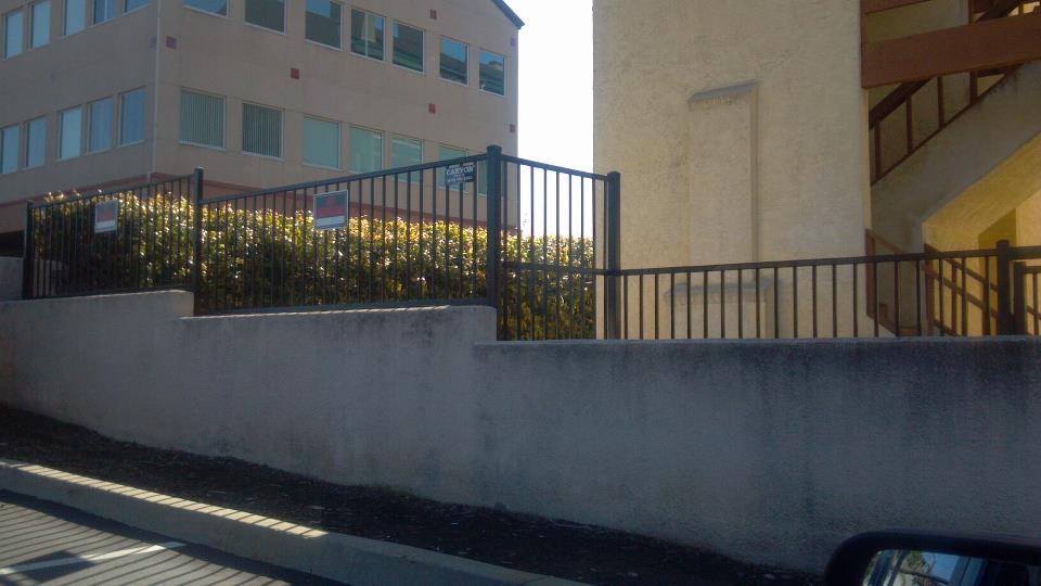 A tall, tan building with windows, a concrete wall, and a black metal fence with bushes in front.