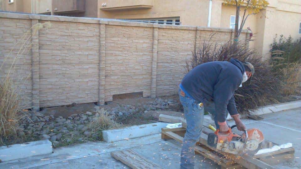 Person cutting wood with a saw near a brown concrete fence and small bushes, in a parking lot.