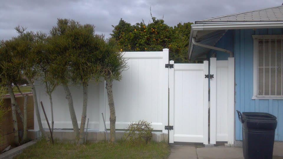 White vinyl fence with gate next to a blue house, small trees in front of the fence, overcast sky.