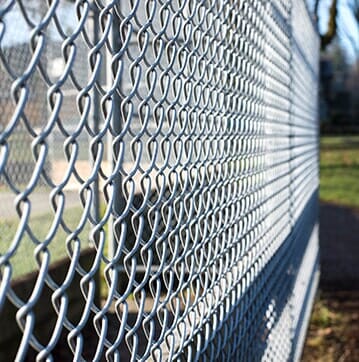 Close-up of a chain-link fence, viewed from an angle. Grey metal links form a pattern against a blurred green background.