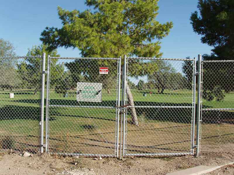 Chain-link fence with double gates blocks access to a park with a tree and sign.