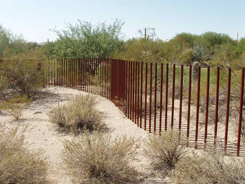 Rusty metal post fence curving through a desert landscape with shrubs and sand.