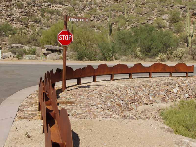 Stop sign at a curve with a decorative, rusty metal fence and a street sign in a desert landscape.