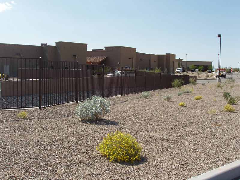 Black fence in front of a tan building with desert landscaping: gravel, shrubs.