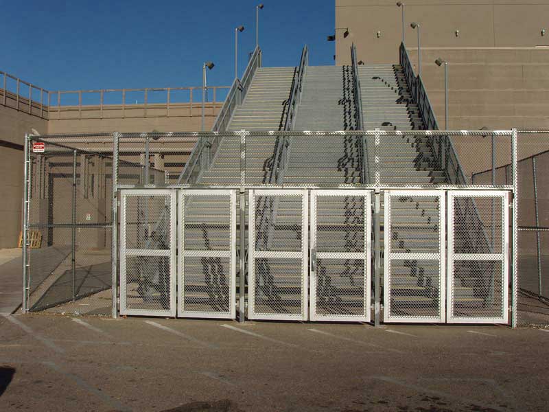 Metal gates leading to concrete stadium stairs, enclosed by fencing, under a bright sky.