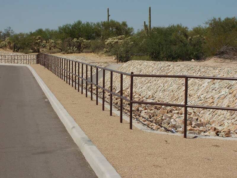 Brown metal railing along road, gravel bed next to rocky slope with desert vegetation in background.