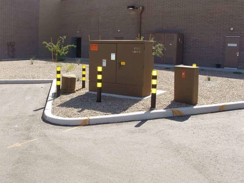 Brown electrical boxes with reflective posts in a gravel area next to a building and a parking lot curb.