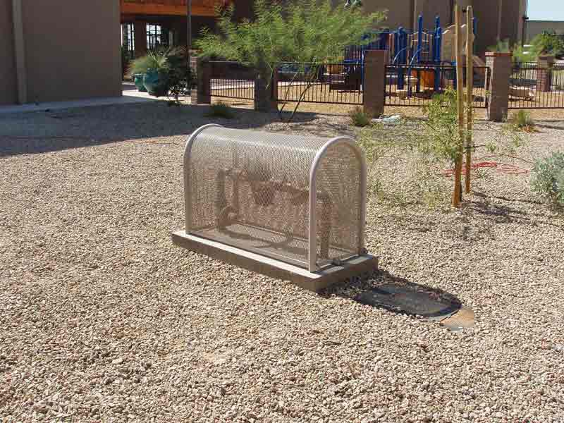 Metal-screened enclosure over utility equipment in a gravel yard near a playground.
