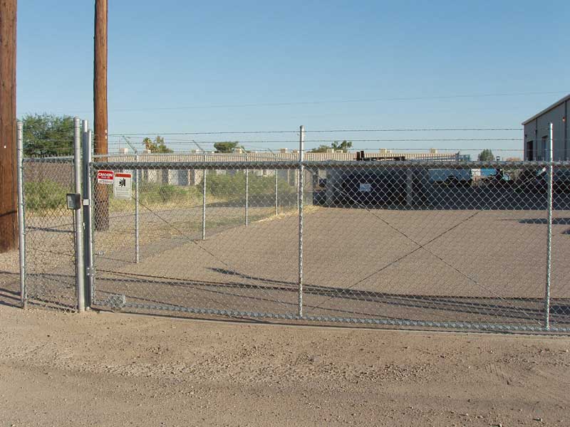 Chain link fence with barbed wire on top, guarding a paved area and industrial buildings.