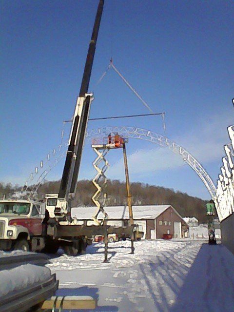 Crane lifting a curved metal structure; workers on a lift. Snow covers the ground near buildings and a truck.