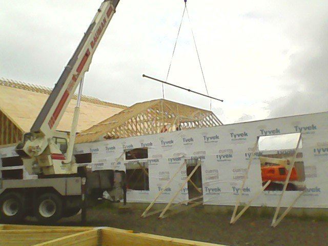 A crane lifts a wall section during house construction; Tyvek wrap, wood frames, and trusses visible.