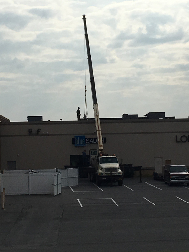 A crane lifting something onto the roof of a Lowe's store. Cloudy sky, parked vehicles, white fence.