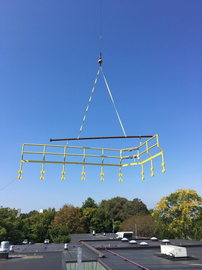 A safety railing suspended in the air by a crane, above a flat roof on a clear day.