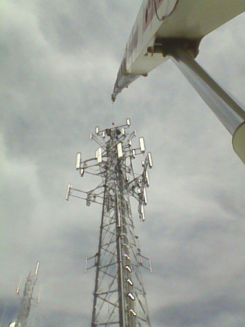 Crane arm reaching towards a tall cell tower under a cloudy sky.