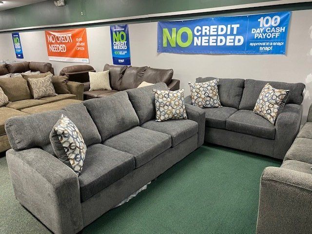 Furniture store display: gray couches and loveseat with patterned pillows, green carpet. 