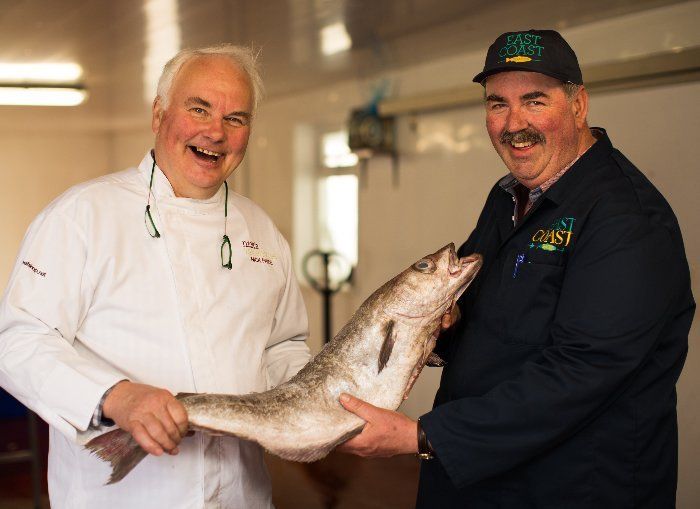 Two men holding a large fish, smiling. One wears a chef's coat, the other, a work uniform. Indoors, possibly a fish market.