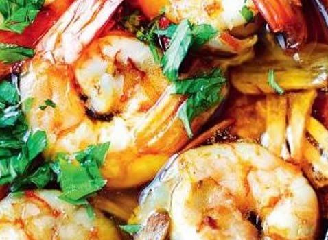 Close-up of cooked shrimp with bright orange shells, green herbs, and cooked vegetables in a reddish sauce.