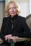 Woman with blonde hair, wearing a black blazer and turtleneck, seated on a chair.