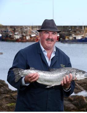 Man holding a large silver fish near a harbor, wearing a hat and navy coat.