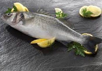 Whole, raw sea bass on a dark slate, garnished with lemon slices and parsley.