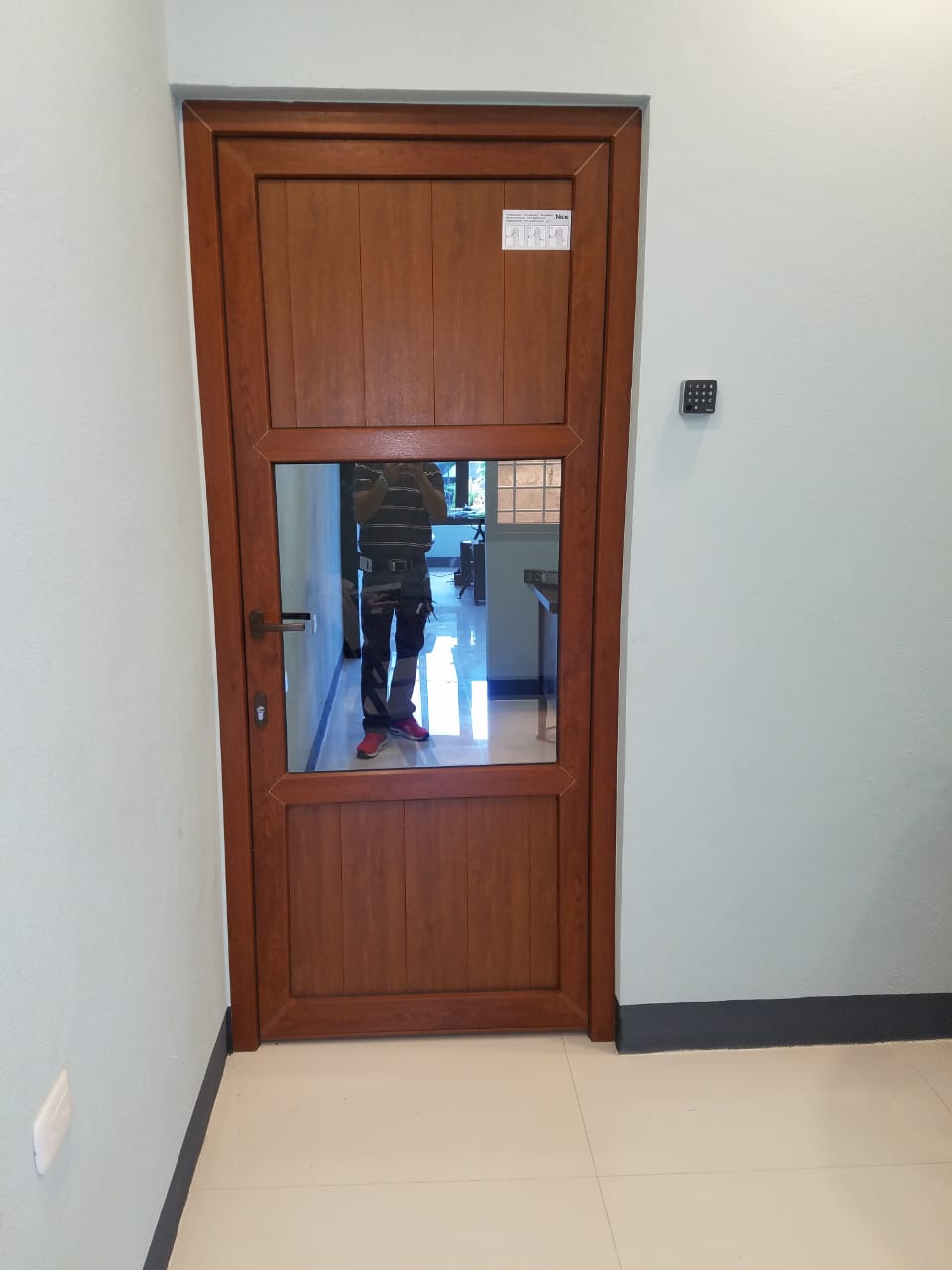 Puerta de madera con una pequeña ventana en un pasillo sencillo, que refleja a una persona y el corredor que hay más allá.