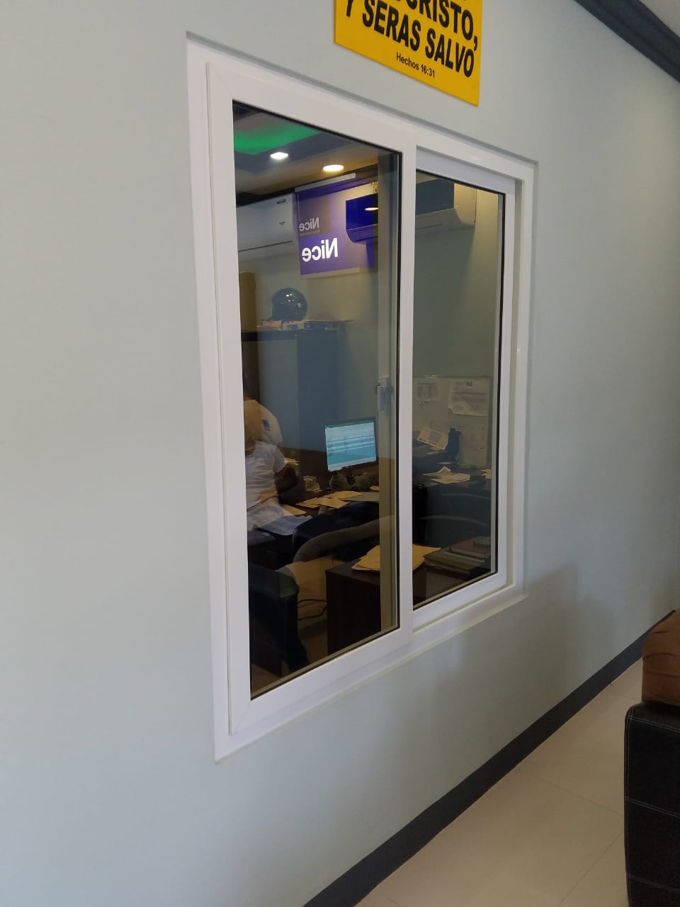 Ventana interior de una oficina con una persona trabajando en un escritorio y un cartel de advertencia