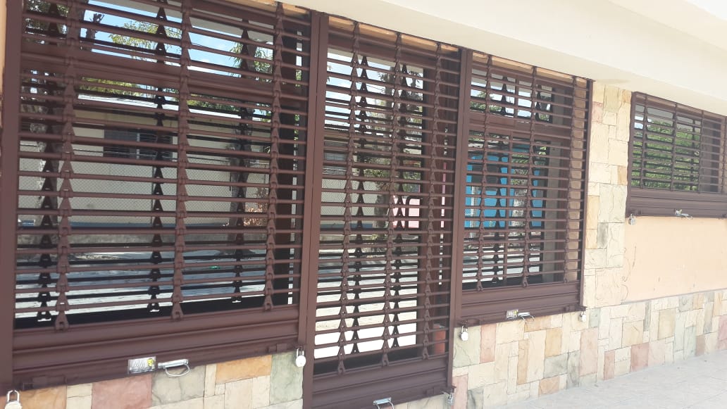 Persianas marrones con listones horizontales sobre una pared exterior beige, encima de una base de piedra.