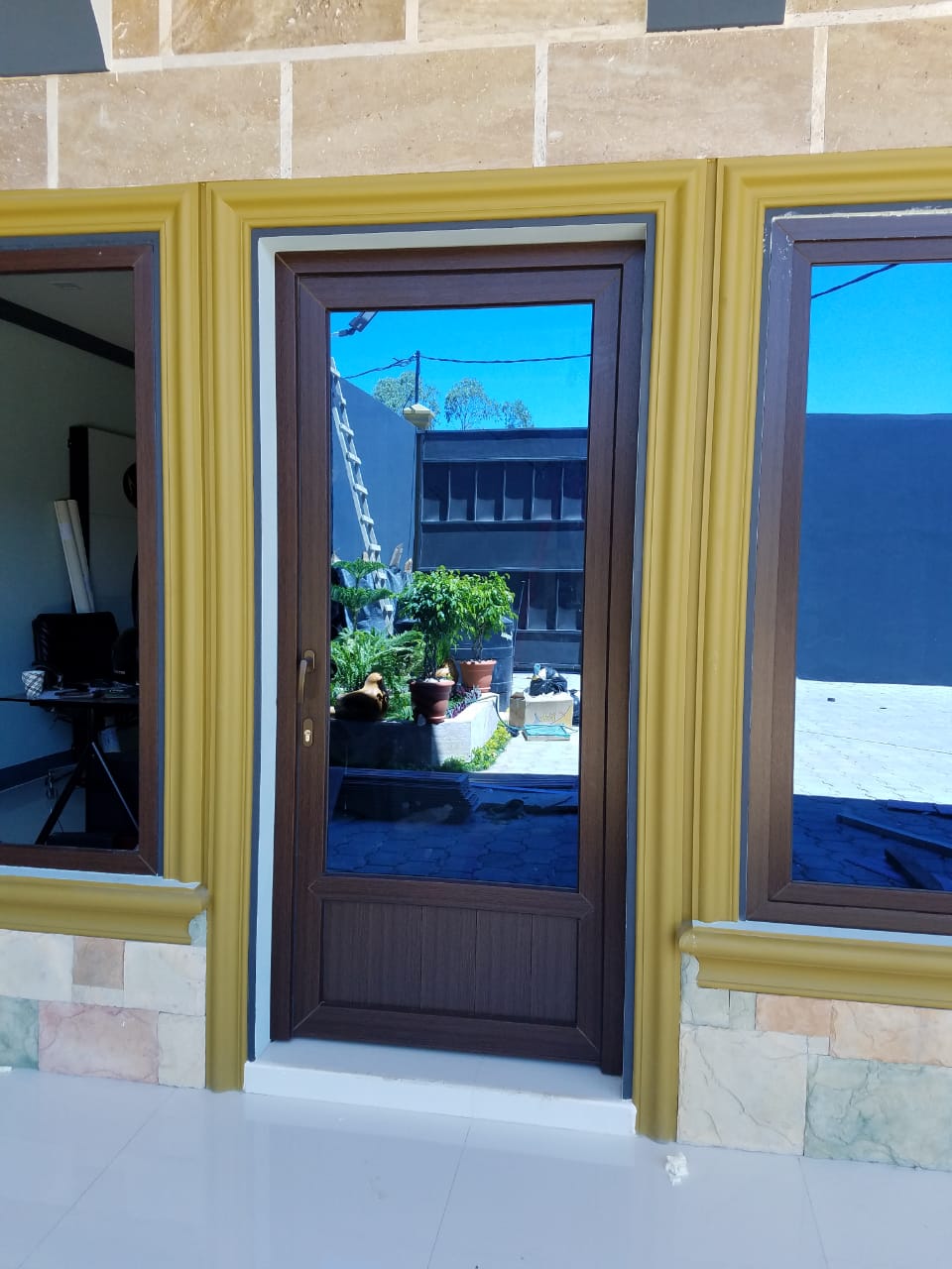 Puerta principal con cristal azul, enmarcada por paredes amarillas y ventanas laterales que reflejan un patio soleado.