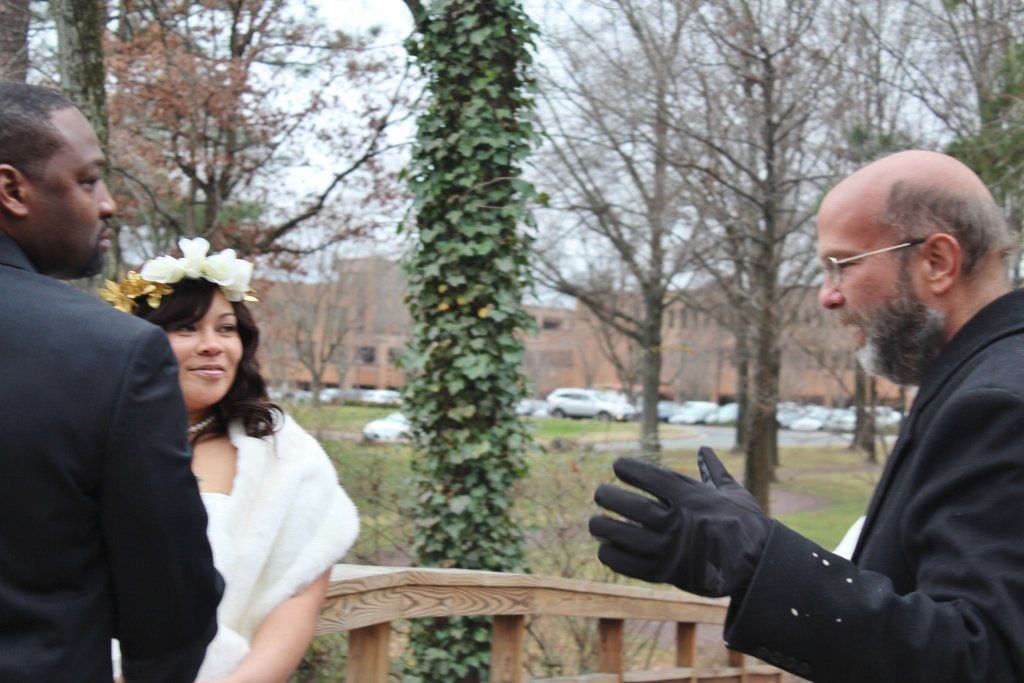 A man in a black suit is talking to a bride and groom in a park.