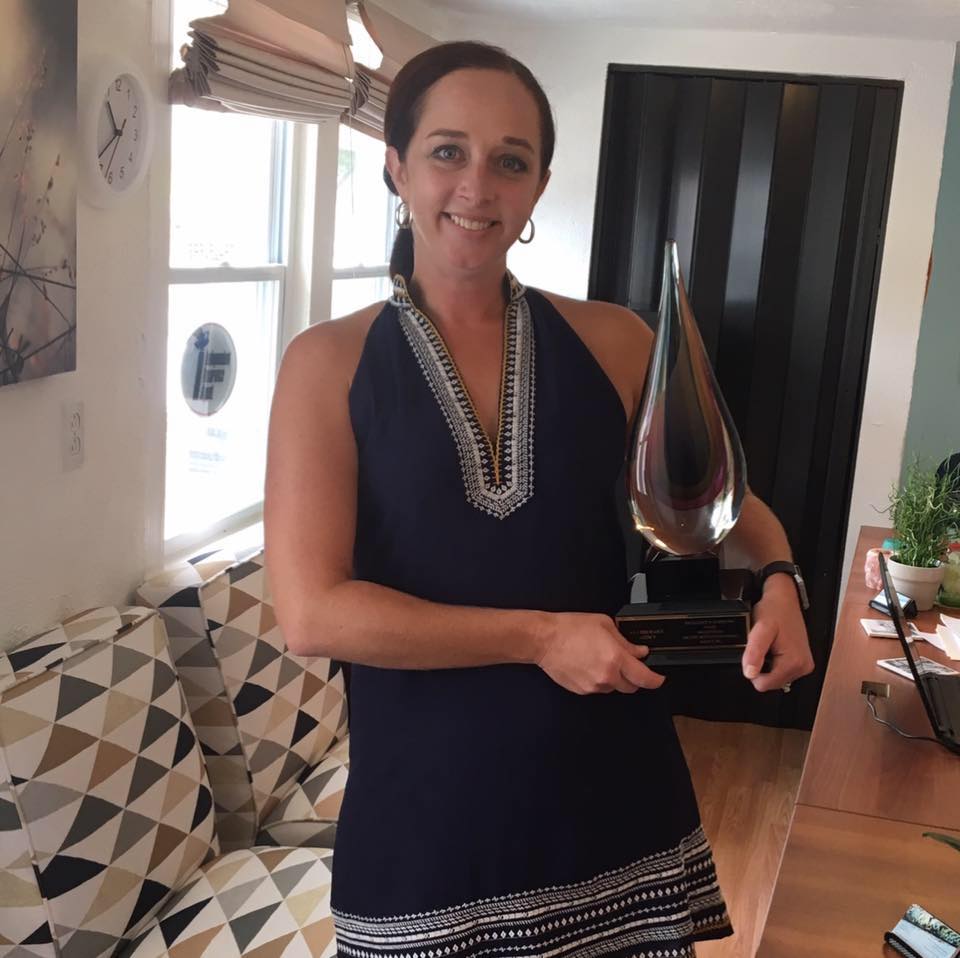 A person in a dark blue, patterned sleeveless dress stands indoors holding a tall, clear, teardrop-shaped glass award.