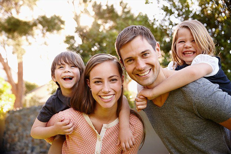 Smiling family of four outdoors in a sunlit park, with two children on their parents' backs